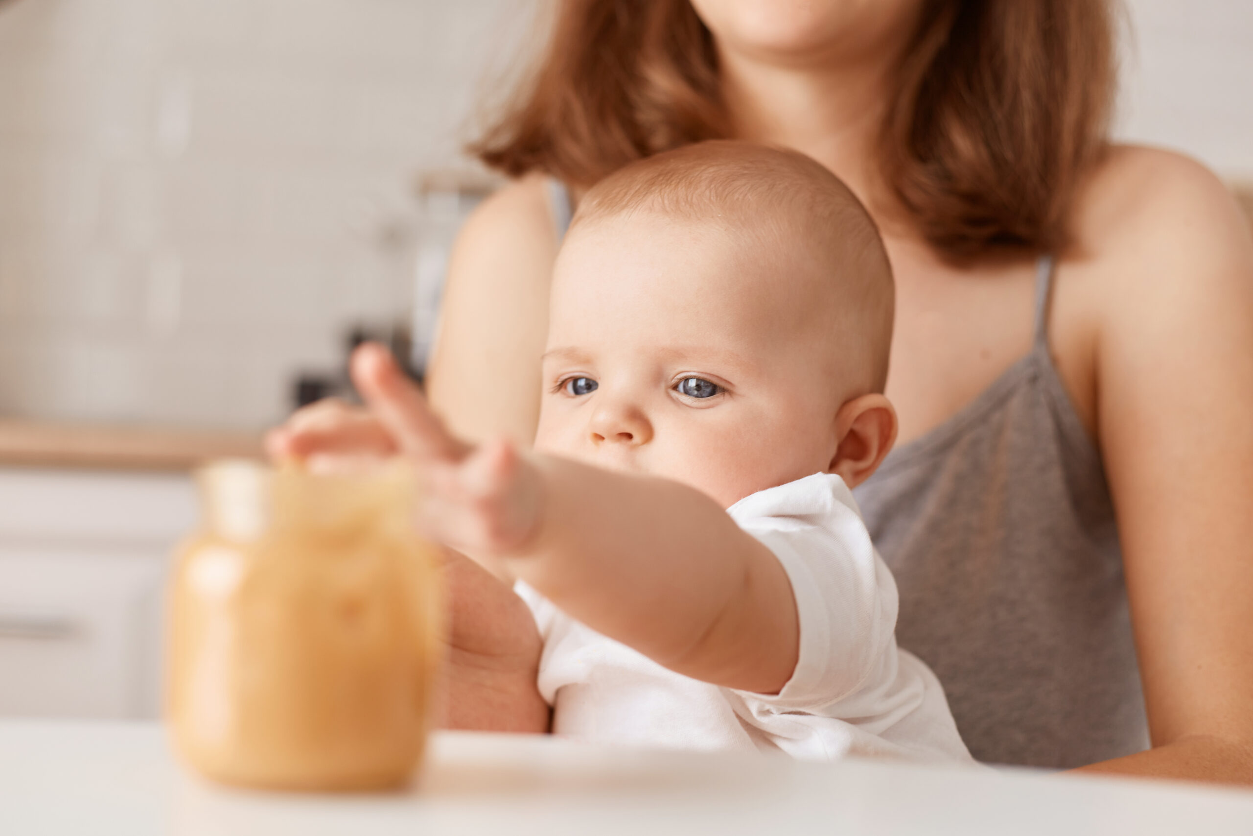 Indoor shot of faceless mother feeding her little infant daughter with vegetable puree, charming toddler baby stretching hand to jar with food, feeding up.